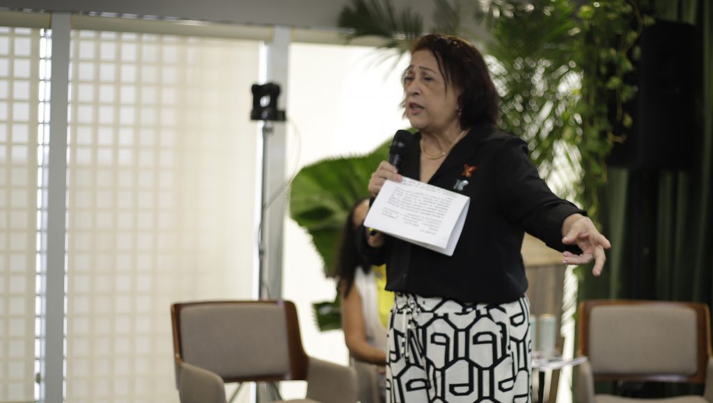 Fotografia registra a professora Ludetana, em cima do palco, falando ao microfone.