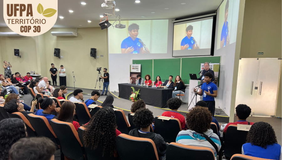 Fotografia registra parte do público em um auditório. Um jovem fala ao microfone e é transmitido nos telões do auditório. Sobre a foto, um selo identifica: UFPA TERRITÓRIO DA COP.
