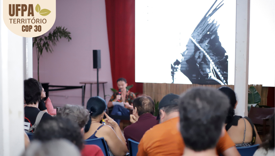 Fotografia registra parte de uma plateia atenta a repodução de um vídeo. Na imagem do telão, um homem de cocar. Sobre a foto, um selo identifica: UFPA TERRITÓRIO DA COP.