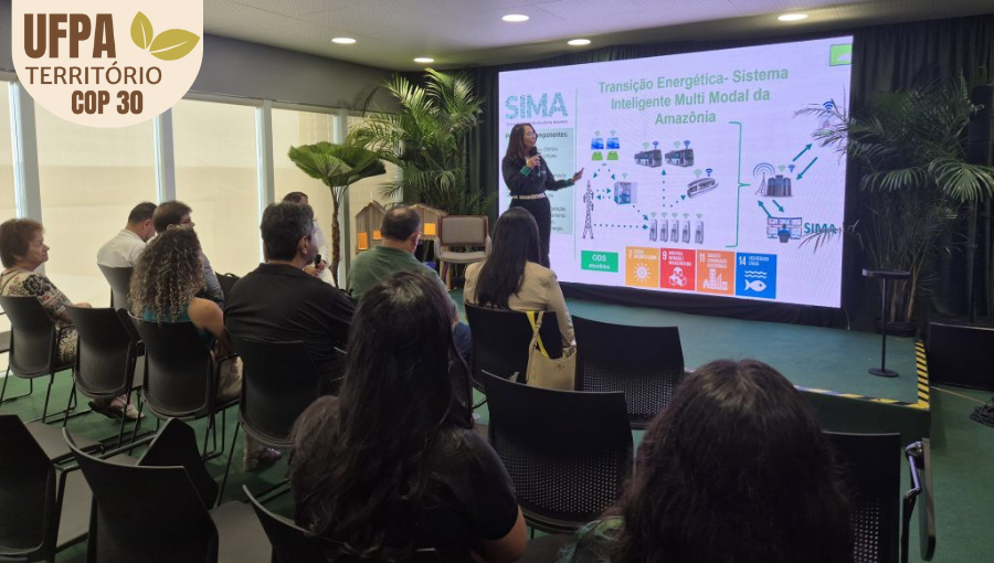 Fotografia registra a professora Emília Tostes, falando ao microfone, durante sua apresentação na Greenzone. Ela é observado por pessoas sentadas na plateia.