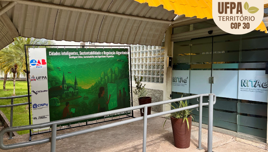Fotografia registra a entrada do prédio do NITAE. Ao lado da porta de entrada há um painel com a seguinte informação: Cidades Inteligentes, Sustentabilidade e Regulação Algorítmica. Sobre a foto, um selo identifica: UFPA TERRITÓRIO DA COP.
