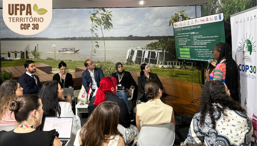 Fotografia registra uma apresentação no stand da UFPA. Uma mulher está em pé, ao lado de uma tela, falando ao microfone. Sobre a foto, um selo identifica: UFPA TERRITÓRIO DA COP.