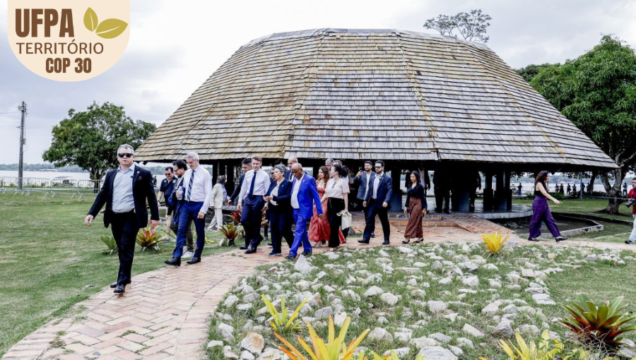Fotografia registra um grupo de pessoas caminhando por uma passarela. Ao fundo, há uma maloca e árvores. Entre as pessoas, o presidente da França, Emmanuel Macron; o reitor da UFPA, Gilmar Pereira da Silva, e a vice-reitora da UFPA, Loiane Prado Verbicaro. Sobre a foto, um selo identifica: UFPA TERRITÓRIO DA COP.