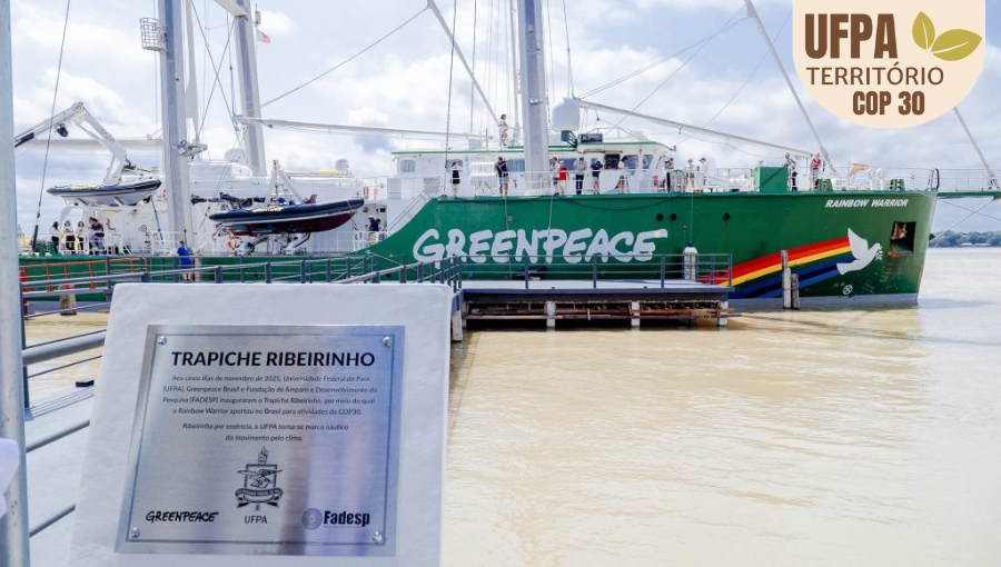 Fotografia registra uma placa de identificação: TRAPICHE RIBEIRINHO, abaixo da informação há o brasão da UFPA e as logomarcas do Greenpeace e da Fadesp. Ao fundo, é possível ver uma grande embarcação na cor verde, identificada com Greenpeace, seguida por um desenho de arco íris e uma pomba branca, com a a descrição "rainbow warrior". Sobre a foto, um selo identifica: UFPA TERRITÓRIO DA COP.