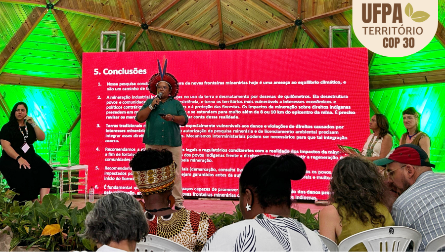 Fotografia de uma mulher indígina, de cocar, falando ao microfone de cima de um palco. Sobre a foto, um selo identifica: UFPA TERRITÓRIO DA COP.
