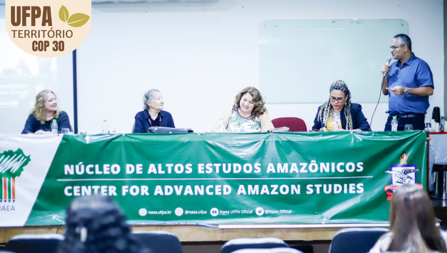 Fotografia registra a mesa de um evento. A mesa está coberta por uma faixa: NÚCLEO DE ALTOS ESTUDOS AMAZÔNICOS. Na mesa, há quatro mulheres sentadas e um homem em pé, falando ao microfone.