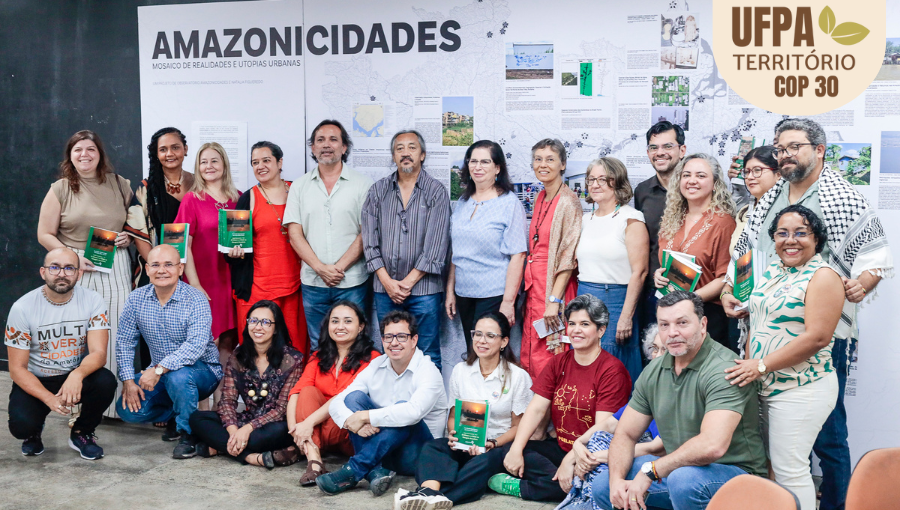 Fotografia posada de um grande grupo de pessoas. Alguns seguram exemplares de um livro. Atrás do grupo, um banner destaca a palavra AMAZONICIDADES. Sobre a foto, um selo identifica: UFPA TERRITÓRIO DA COP.