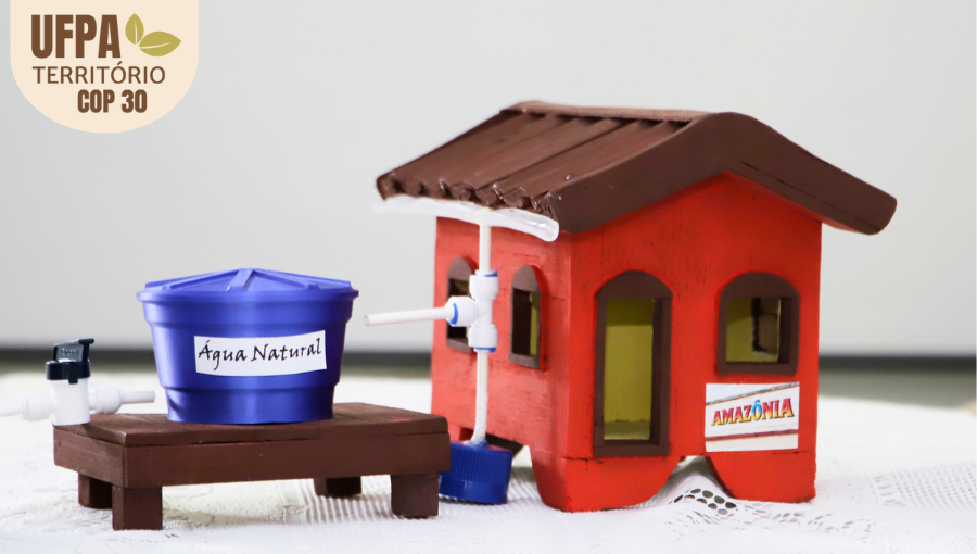 Fotografia registra uma miniatura de uma casa de palafitas ao lado de uma caixa d'água. Sobre a foto, um selo identifica: UFPA TERRITÓRIO DA COP.