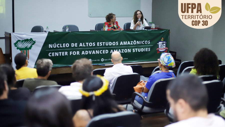 Fotografia registra a mesa de um evento. A mesa está coberta por uma faixa: NÚCLEO DE ALTOS ESTUDOS AMAZÔNICOS. Na mesa, há duas mulheres sentadas. Uma delas fala ao microfone.