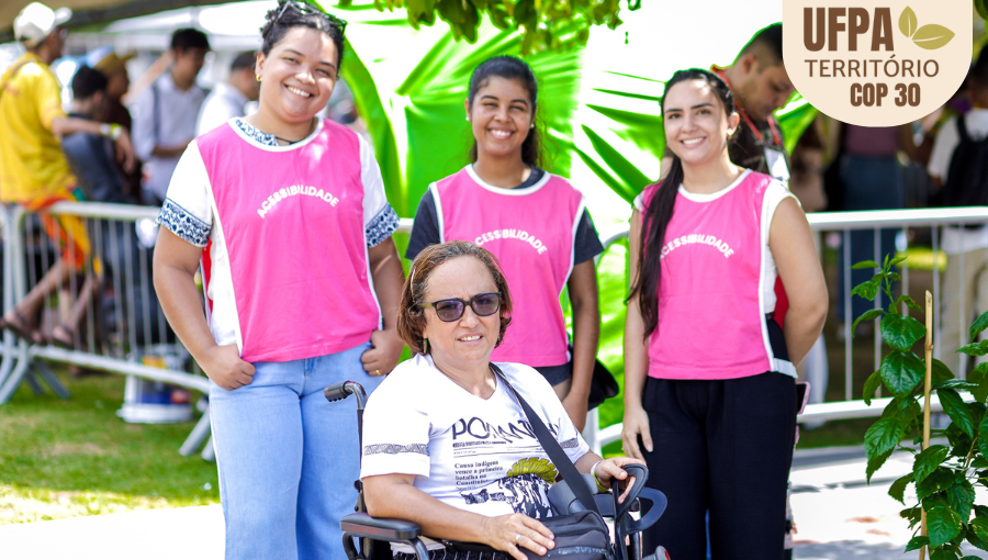 Fotografia registra quatro pessoas. Três delas estão em pé, identificadas com coletes rosa de monitoras. A quarta pessoa, está em uma cadeira de rodas na frente das três mulheres. Sobre a foto, um selo identifica: UFPA TERRITÓRIO DA COP.