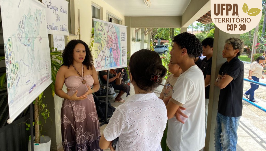 Fotografia registra uma mulher apresentando um painel, em um ambiente aberto, para um grupo de jovens.