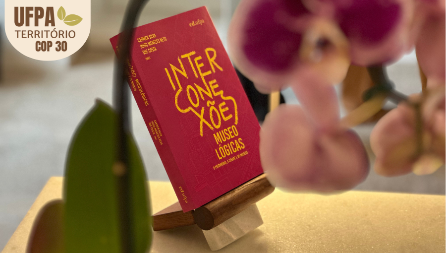Fotografia registra, em segundo plano, a capa do livro Interconexões. Eles está sobre um suporto. Em primeira plano, uma orquídea. Sobre a foto, um selo identifica: UFPA TERRITÓRIO DA COP.