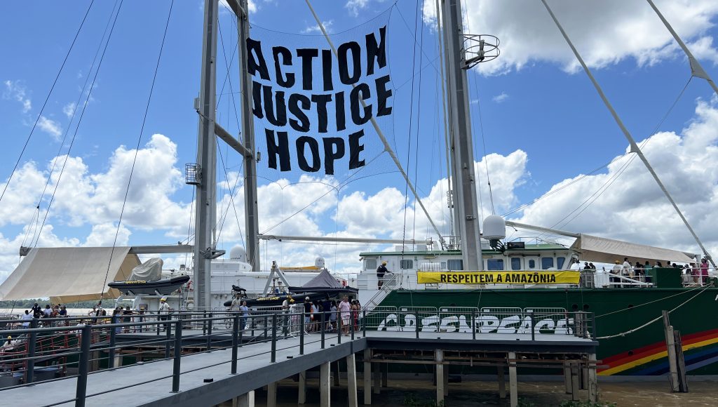 Fotografia registra uma grande embarcação ancorada em um pier. Uma tela, no topo da embarcação, destaca a mensagem: ACTION JUSTICE HOPE.