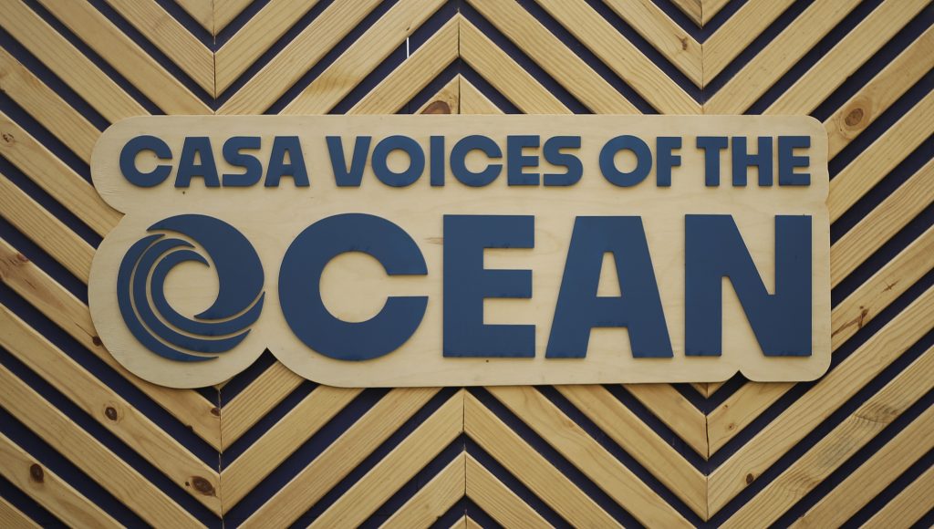 Painel de madeira destaca um letreiro: CASA VOICES OF THE OCEAN.