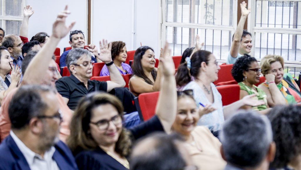 Fotografia de parte da plateia de um auditório. Algumas pessoas sorriem e levantam os braços.