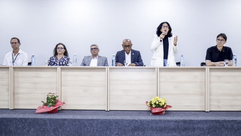 Fotografia registra a mesa de um evento. Nela estão presentes o reitor da UFPA, Gilmar Pereira da Silva; a pró-reitora de Pesquisa e Pós-graduação, Iracilda Sampaio. Ela está em pé, falando ao microfone.