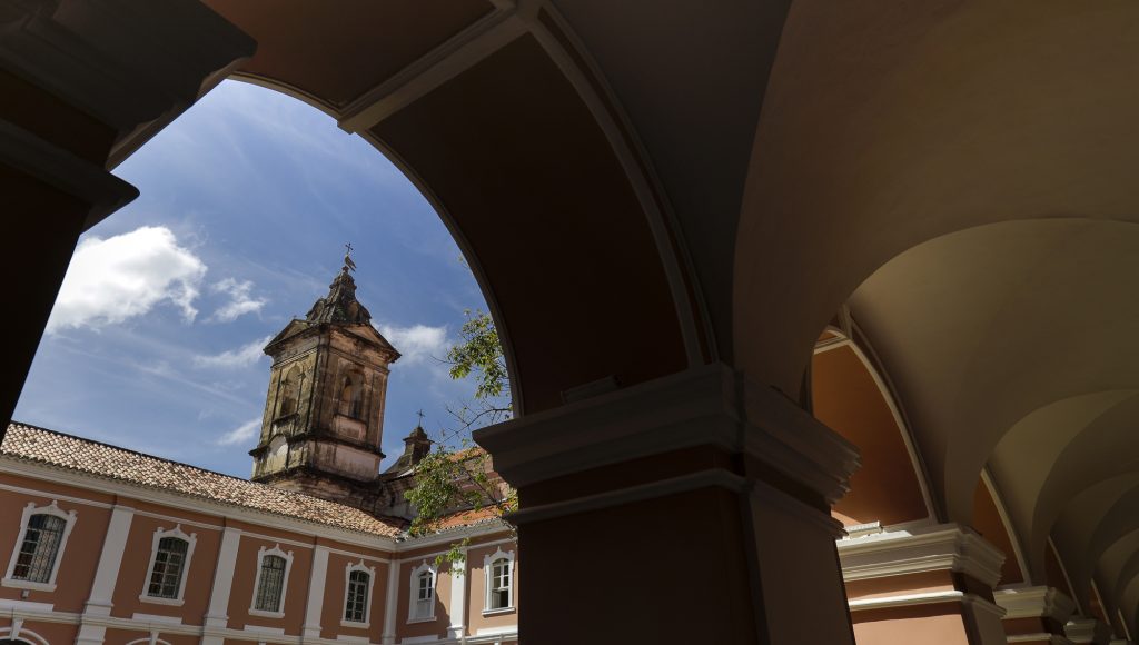 Fotografia mostra parte interna da arquitetura do Convento Mercedários. A estrutura tem predominância da cor rosa com detalhes em branco. Ao fundo, tem-se a torre de uma igreja ainda não restaurada.