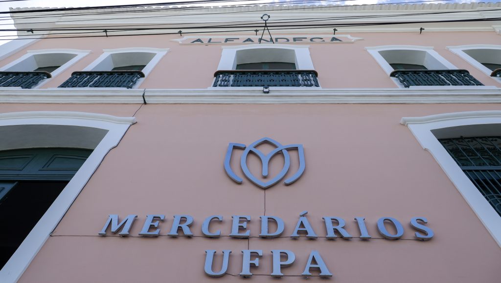 Fotografia registra a fachada de um prédio de parede rosa. Em destaque, um letreiro informa "MERCEDÁRIOS UFPA", bem no topo da imagem, um outro letreiro menor informa "ALFÂNDEGA".