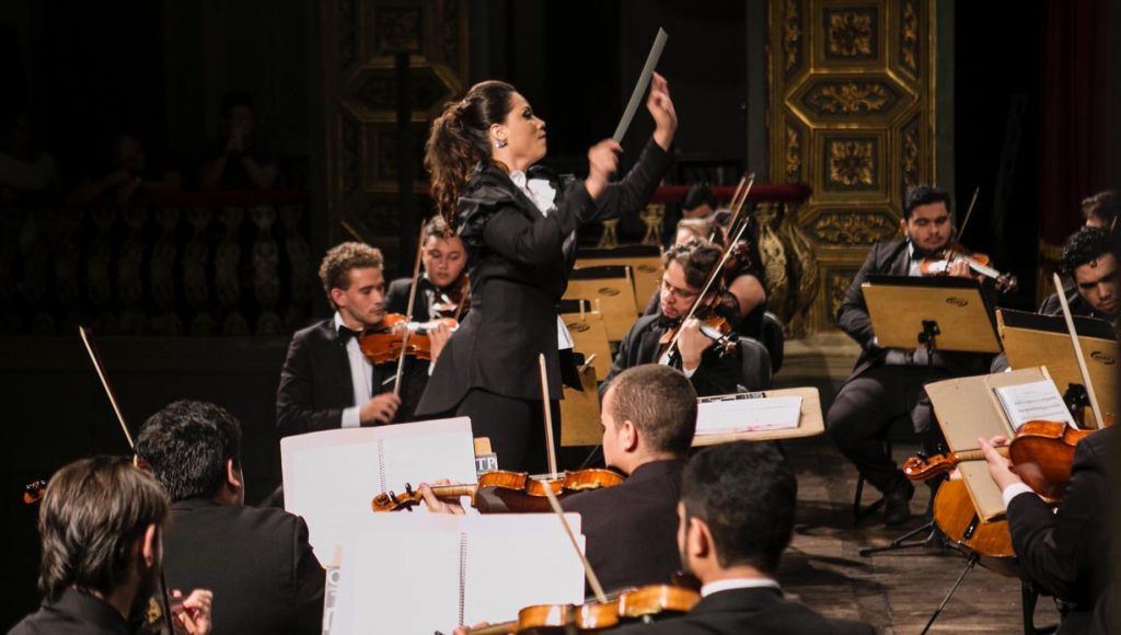 Fotografia registra a professora Cibelle Donza, meio a uma orquestra, regendo o grupo.