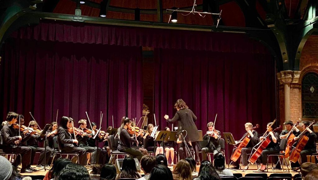 Fotografia registra a professora Cibelle Donza, meio a uma orquestra, regendo o grupo. Ela está de costa para a plateia.