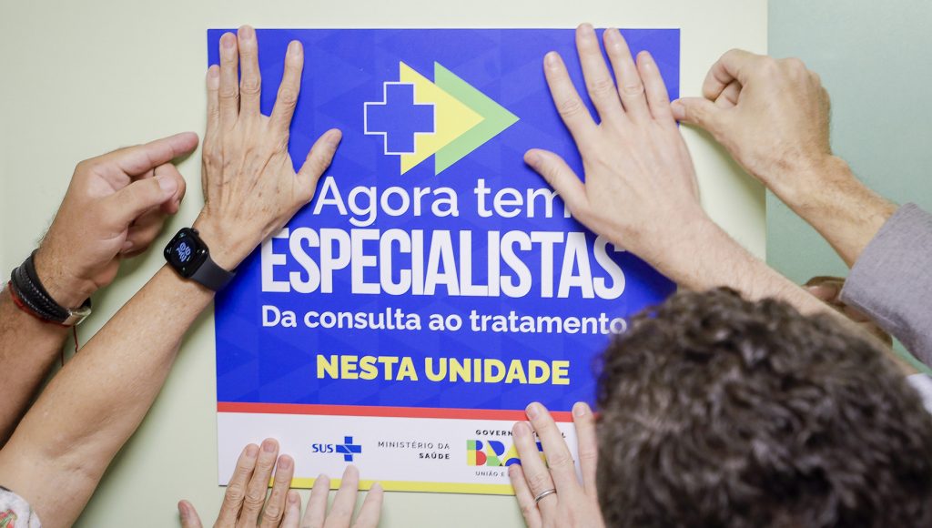 Fotografia registra mãos de diferentes pessoas segurando uma placa que informa: AGORA TEM ESPECILISTAS - DA CONSULTA AO TRATAMENTO - NESTA UNIDADE.