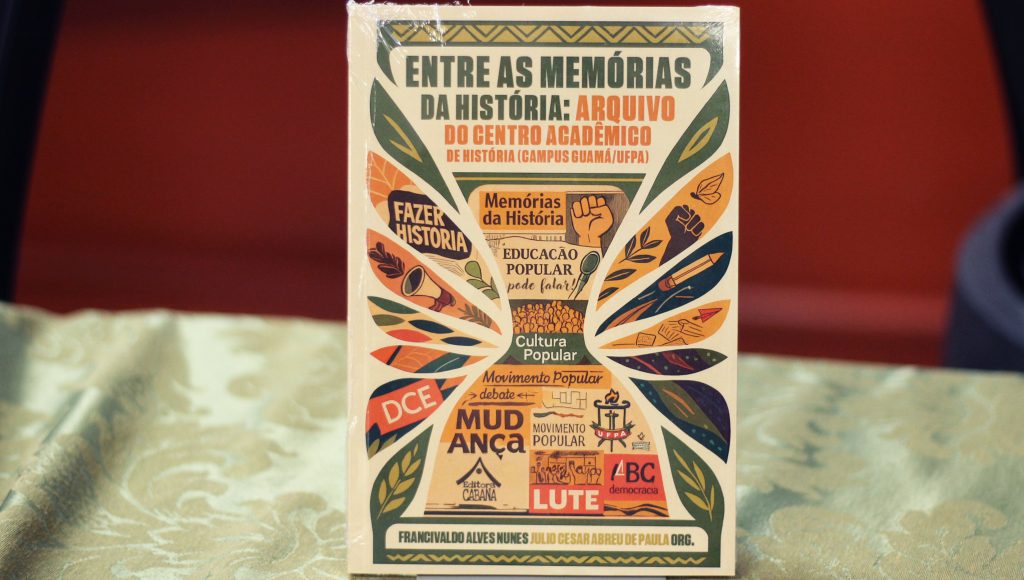 Fotografia registra a capa de um livro. Entre quadro coloridos e um desenho que remete a uma ampulheta, a capa destaca o seguinte texto: Entre as memórias da história: Arquivo do Centro Acadêmico de História.