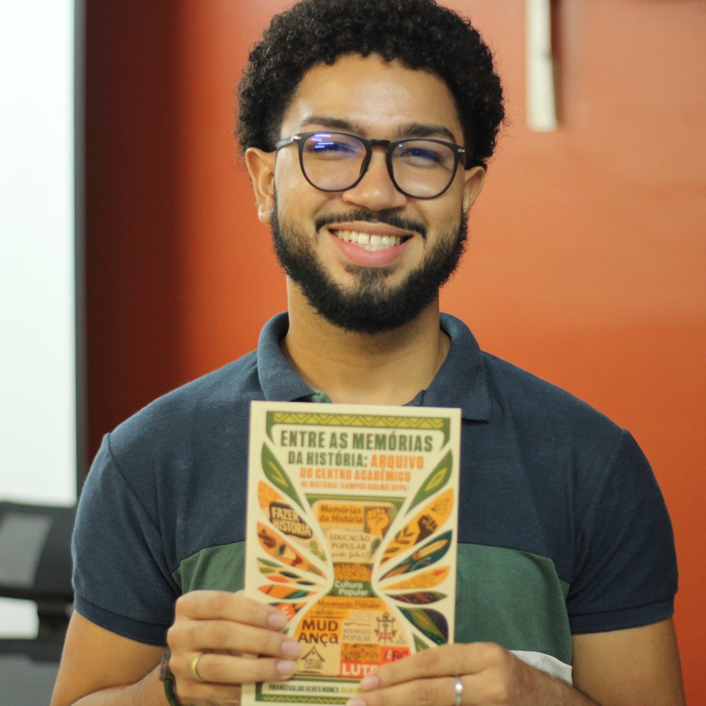 Fotografia posada registra o estudante Lucas segundo um exemplar do livro em mãos. Lucas é um homem negro, de cabelos pretos curtos, usa óculos de armação redonda preta e sorri para a foto.