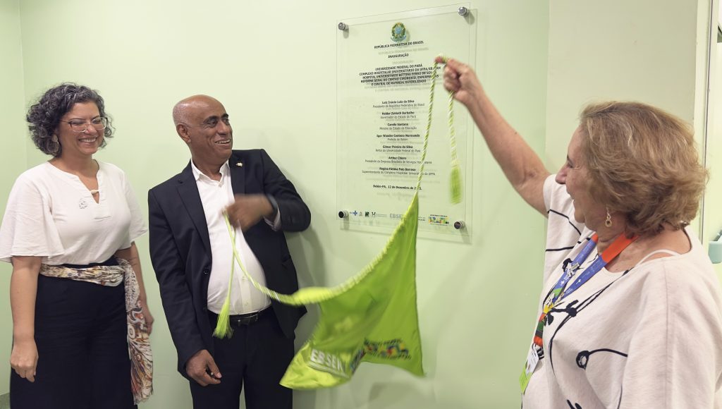 Fotografia registra o momento de descerramento de uma placa de inauguração. Na foto, é possível observar o reitor da UFPA, Gilmar Pereira da Silva segurando um lado da faixa, e a superintendente dos hospitais, Regina Feio, segurando o outro lado da faixa.