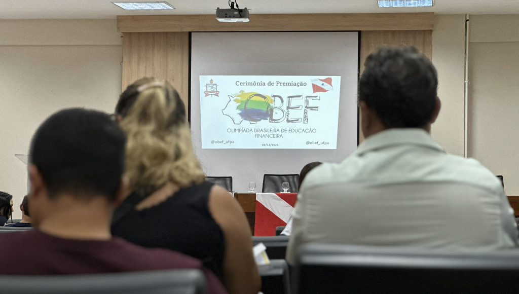 Fotografia registra, em primeiro plano, algumas pessoas sentadas. Ao fundo, um telão destaca a seguinte informação: CERIMÔNIA DE PREMIAÇÃO DA OLIMPÍADA BRASILEIRA DE EDUCAÇÃO FINANCEIRA - OBEF.
