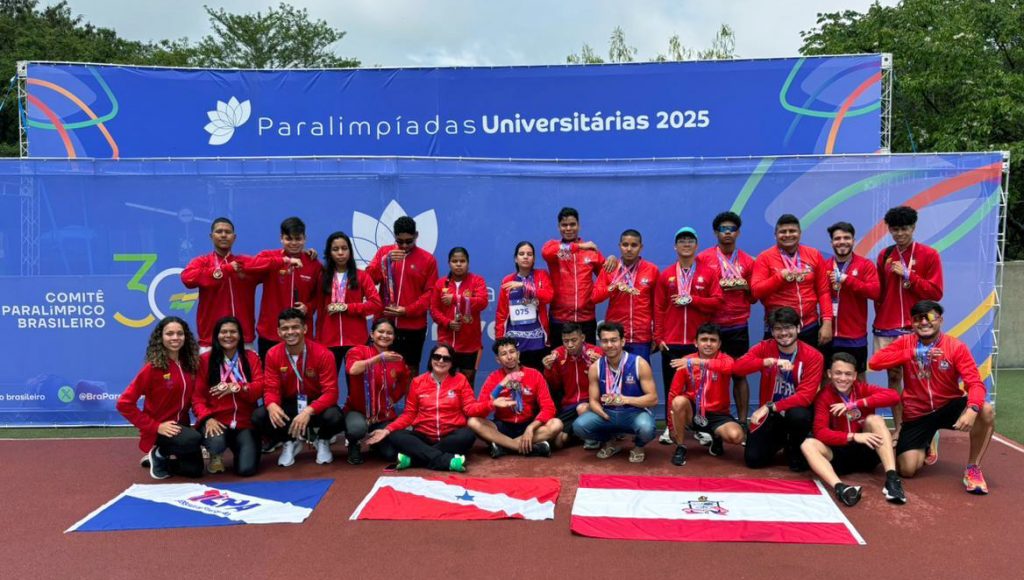 Fotografia posada de um grupo de pessoas. Eles estão dispostos na frente de um painel que destaca a informação: PARALIMÍADAS UNIVERSITÁRIAS 2025. Várias pessoas possuem medalhas nos pescoço e alguns possuem bengalas brancas. Na frente do grupo, no chão, estão dispostas três bandeiras: da UEPA, do Pará e da UFPA.