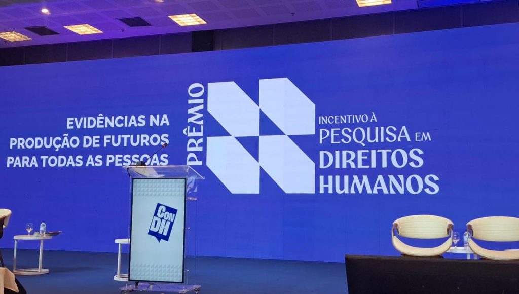 Fotografia registro um telão azul com a seguinte informação: evidências na produção de futuros para toas as pessoas - 1º Prêmio de Incentivo à Pesquisa em Direitos Humanos.
