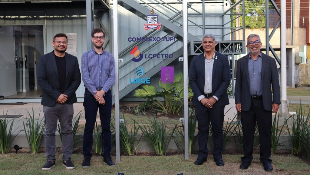 Fotografia posada de um grupo de quatro pessoas. Eles estão postados ao lado de uma placa que identifique: COMPLEXO TUPI, seguido pelo brasão de UFPA e outras logomarcas.