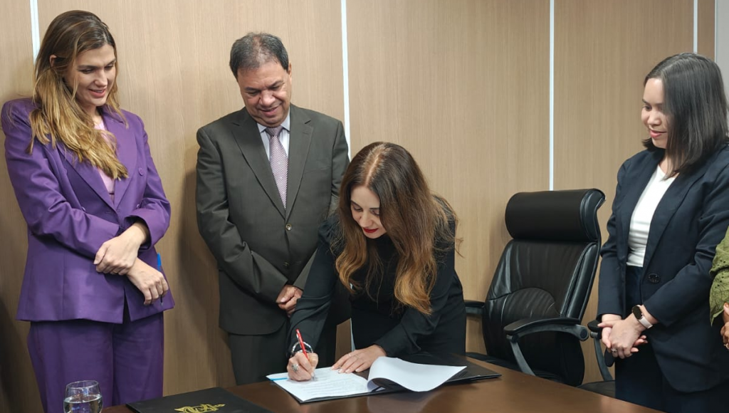 Fotografia registra a vice-reitora da UFPA, Loiane Prado Verbicaro, assinando um documento disposto em uma mesa de madeira. Ela é observada por outras três pessoas.