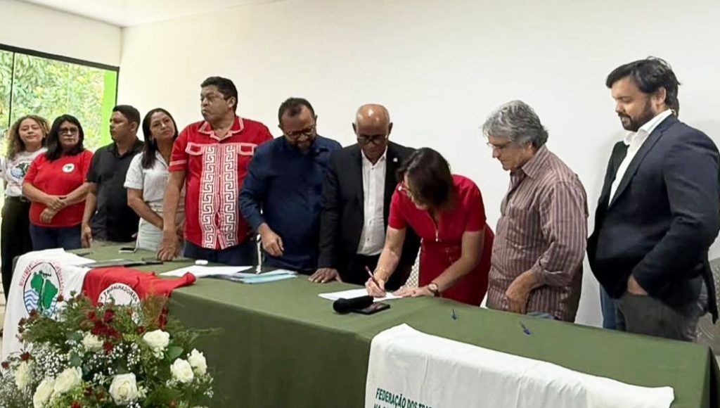 Fotografia registra um grupo de pessoas atrás de uma mesa. Uma mulher assina um documento em cima da mesa.