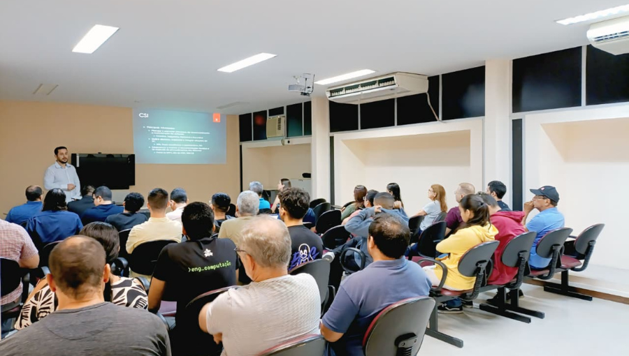 Fotografia mostra público sentado de costas. Ao fundo, um homem está de pé e de frente para a plateia.
