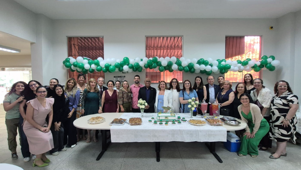 Fotografia posada de um grande grupo de pessoas ao redor de uma mesa com bolo e doces. Ao fundo, há balões nas cores verde e branco decorando o espaço.