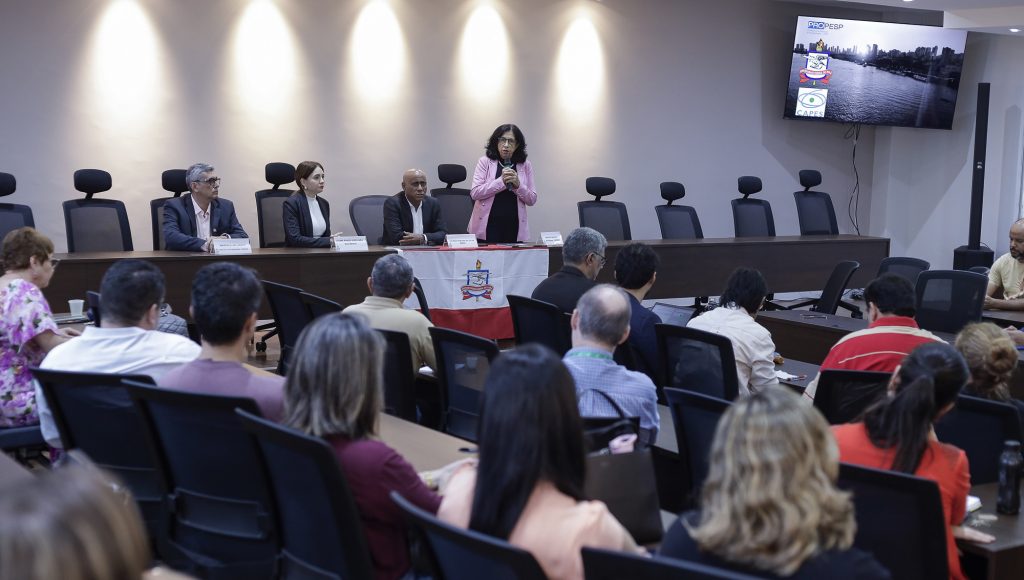 Fotografia registra parte da plateia do auditório. Ao fundo, na mesa do evento, o reitor da UFPA, Gilmar Pereira da Silva, a vice-reitora da UFPA, Loiane Prado Verbicaro, a pró-reitora de Pesquisa e Pós-Graduação, Iracilda Sampaio, e o professor diretor de Pós-Graduação da Propesp, Marcelo Vallinoto.