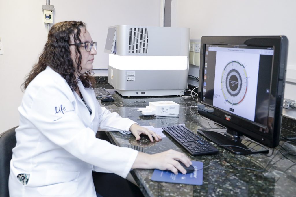 Fotografia mostra mulher sentada na frente de um tela de computador. Ela usa jaleco e segura o mouse com uma luva.