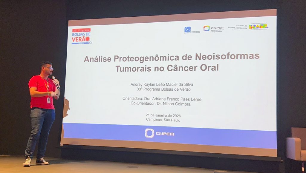 Fotografia registra um homem, falando ao microfone, próximo a um telão. No telão, há a seguinte informação: Análise Proteogenômica de Neoisoformes Tumorais no Câncer Oral - ANDREY KAYLAN LEÃO MACIEL DA SILVA - 33º PROGRAMA BOLSAS DE VERÃO - ORIENTADORA ADRIANA FRANCO PAES LEME - CO-ORIENTADOR DR. NILSON COIMBRA - 21 DE JANEIRO DE 2026 - CAMPINAS - SÃO PAULO.