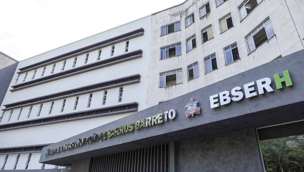Fotografia registra parte da fachado do Hospital Universitário João de Barros Barreto. O letreiro com o nome do hospital é seguido pelo brasão da UFPA e por um letreiro da Ebserh.