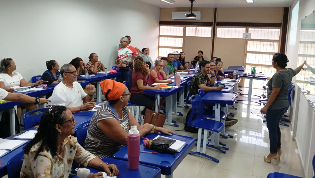 Fotografia registra uma sala de aula. Nela, algumas pessoas estão sentadas em carteiras azuis, atentas à uma pessoa que gesticula em frente a um quadro branco.