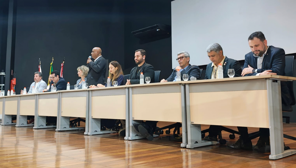 Fotografia registra a mesa de um evento. No centro da mesa, o reitor, Gilmar Pereira da Silva, em pé, fala ao microfone.