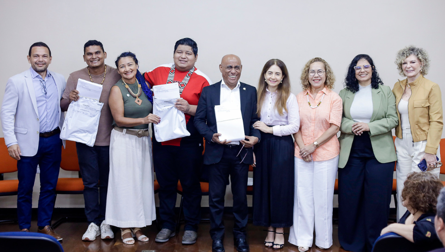 Fotografia de um grupo de pessoas de pé. Eles seguram envelopes de plástico com folhas de papel à mostra.