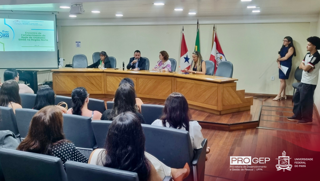 Fotografia registra a mesa de um evento. Ao lado da mesa, em um telão, a palavra SIASS.