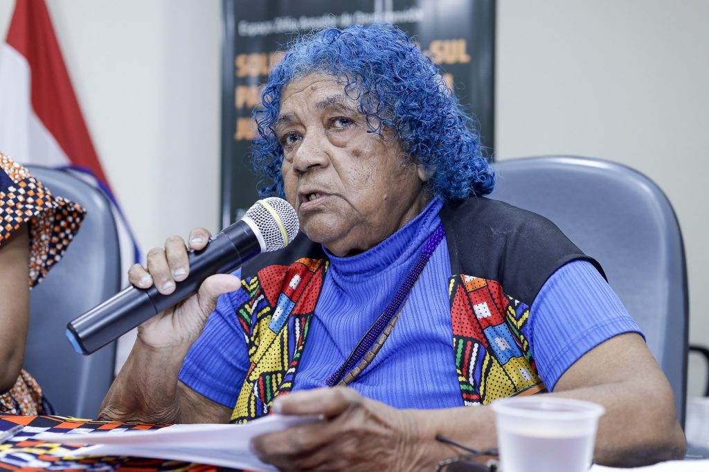 Fotografia mostra Nilma Bentes. Mulher negra, tem cabelos crespos de cor azul e fala ao microfone.