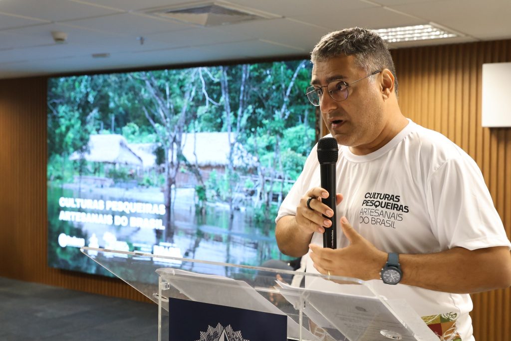 Fotografia do professor Flávio Barros. Homem pardo, grisalho. Ele usa blusa branca e fala ao microfone. 