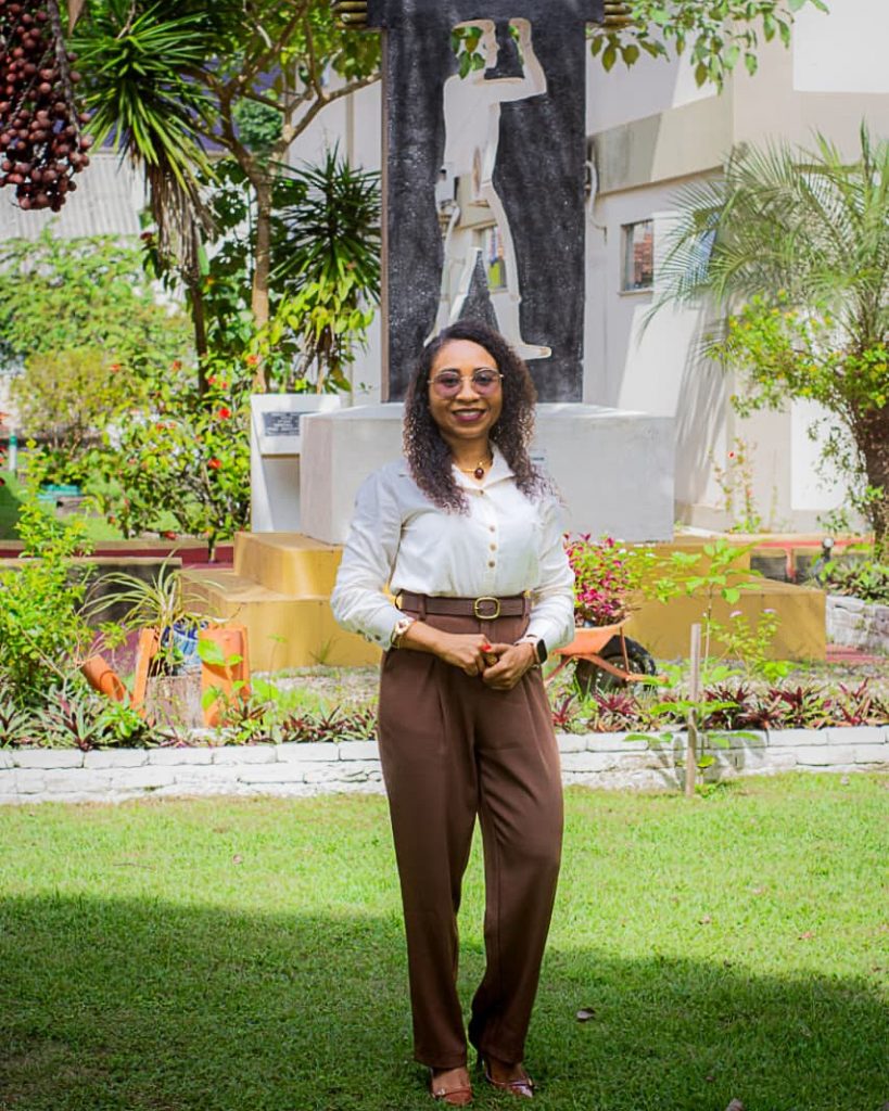 Fotografia posada de Ladyana dos Santos Lobato. Ela é uma mulher negra, de cabelos cacheados na altura do ombro. ELA USA BLUSA BRANCA E CALÇA MARROM. Ela está em uma área verde com um monumento ao fundo.