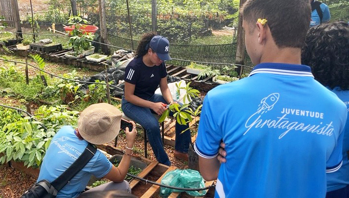 Fotografia registra Ayla em um ambiente que se assemelha a uma horata. Ela está sentada, manuseado uma planta com um alicate. A ação é registrada por uma outra pessoa que fotografa com um celular. Em primeiro plano, um jovem observa o ambiente de costa para a foto. Na sua camisa, a seguinte informação: JUVENTUDE PROTAGONISTA.