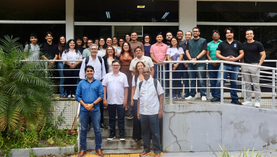 Fotografia mostra grupo de muitas pessoas em pé posando para a fotografia. São homens e mulheres. Eles estão na frente de um prédio.