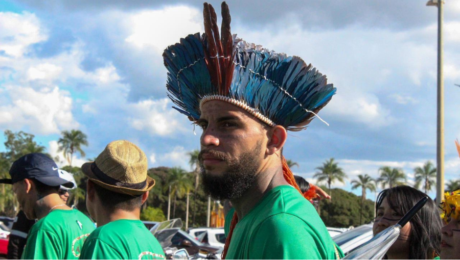 Fotografia mostra homem com cocar de penas azuis e vermelhas. Ele veste camisa camisa verde.
